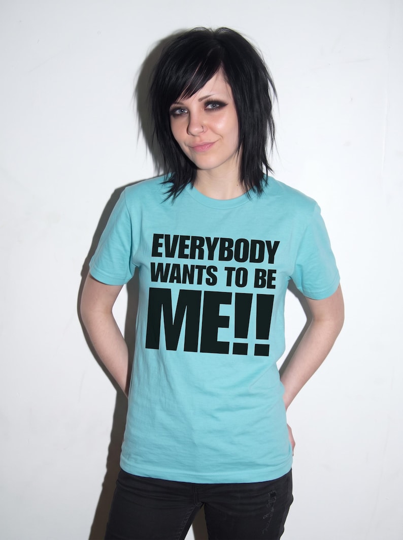 Puede incluir: Una camiseta de manga corta de color azul claro con el texto negro "EVERYBODY WANTS TO BE ME!!" impreso en una fuente negrita sin remates en el pecho. La prenda tiene un cuello redondo cl&aacute;sico y un dobladillo recto.