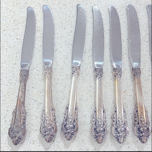 Puede incluir: Un juego de seis cuchillos de mantequilla plateados. Cada cuchillo presenta una hoja curva y un mango ornamentado con un diseño floral. Los cuchillos están dispuestos en fila, mostrando su elegante diseño, ideal para cenas formales.