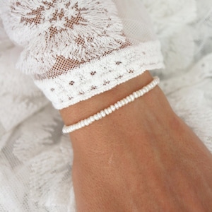 Könnte beinhalten: Ein zartes weißes Perlenarmband am Handgelenk einer Person. Das Armband besteht aus kleinen, gleichmäßig verteilten Perlen.