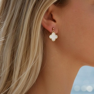 Peut inclure: Boucles d'oreilles dorées en forme de trèfle à quatre feuilles avec un centre blanc, portées à l'oreille d'une personne. Les boucles d'oreilles ont un petit clou doré en haut. L'arrière-plan est une vue floue de l'océan.