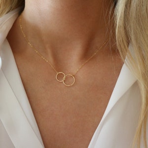 Könnte beinhalten: Eine zarte goldene Halskette mit zwei ineinandergreifenden Kreisen. Die Kreise sind durch eine feine Kette verbunden, wodurch ein minimalistisches und elegantes Design entsteht. Die Halskette wird vor einem weißen Hintergrund gezeigt.