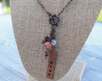 May include: A long copper chain necklace with a charm featuring a cluster of pink and brown stones, a clear crystal, and a copper tag with the word "MTNS" engraved on it.