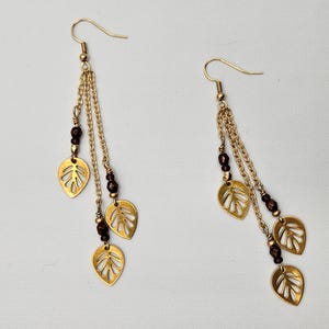May include: A pair of gold-colored dangle earrings. Each earring features a gold hook, chains, leaf-shaped charms, and dark red beads. The earrings are displayed against a white background.