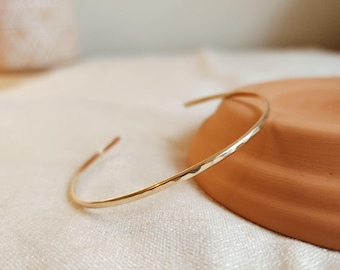 May include: A thin, gold-colored metal cuff bracelet with a hammered, textured surface. The bracelet is circular in shape with an open gap at one end, featuring a polished, reflective finish that catches the light along its curved, slender band.