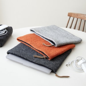 May include: Three felt laptop sleeves stacked on top of each other. The top sleeve is grey, the middle sleeve is orange, and the bottom sleeve is dark grey. Each sleeve has a zipper closure and a leather pull tab.