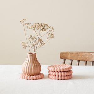 Puede incluir: Un juego de cuatro posavasos de fieltro rosa con un dise&ntilde;o texturizado con forma de burbujas. Los posavasos est&aacute;n apilados uno encima del otro y est&aacute;n sobre un mantel blanco. Un jarr&oacute;n con flores secas est&aacute; en el fondo.