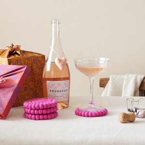 Puede incluir: Tres posavasos de fieltro rosa con una copa de prosecco rosado y una botella de prosecco sobre un mantel blanco. La botella tiene un charm de coraz&oacute;n rosa.