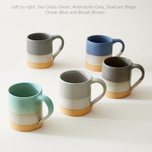 Puede incluir: Cinco tazas de cerámica con esmaltes de diferentes colores. Las tazas están dispuestas en semicírculo sobre una superficie blanca. Las tazas son todas del mismo tamaño y forma, y tienen un diseño simple y moderno. Los colores de las tazas son verde vidrio de mar, gris antracita, beige espuma de mar, azul océano y marrón basalto.