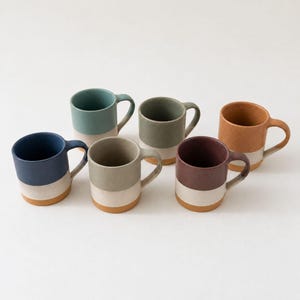 May include: A set of six ceramic mugs, each featuring a two-tone design with a colored upper section and a solid tan base. The mugs are arranged in a group and come in various colors including blue, teal, sage green, olive, maroon, and terracotta.