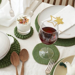Puede incluir: Una mesa puesta con posavasos de fieltro verde, una vela blanca en un tarro de vidrio, una copa de vino tinto y una servilleta blanca con adornos de estrellas doradas.