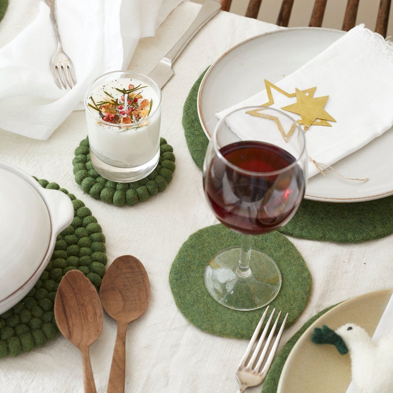 Puede incluir: Una mesa puesta con posavasos de fieltro verde, una vela blanca en un tarro de vidrio, una copa de vino tinto y una servilleta blanca con adornos de estrellas doradas.