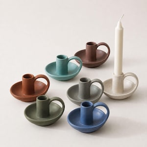 May include: A collection of seven ceramic candle holders in various colours including terracotta, teal, brown, grey, olive green, blue, and speckled cream. Each holder features a circular base, a central cylindrical socket, and a small side handle.