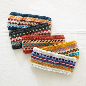 May include: Four knitted headbands with colourful geometric patterns. The headbands are stacked on top of each other, with the top headband being a light grey with orange, blue, and red stripes. The headband below it is a mustard yellow with purple, blue, and green stripes. The next headband is a dark red with blue, green, and yellow stripes. The bottom headband is a dark grey with green, red, and blue stripes.