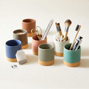 May include: A collection of seven cylindrical ceramic cups in various matte colors including blue, terracotta, sage green, and speckled white. The cups feature a distinct unglazed tan base. They hold items like scissors, pencils, brushes, and a tea bag.