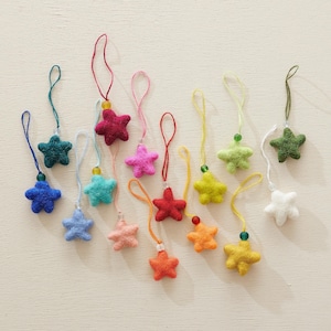Pode incluir: Uma coleção de doze pequenos ornamentos em forma de estrela de cinco pontas feitos de lã feltrada. Cada estrela apresenta uma cor sólida vibrante, incluindo vermelho, azul, verde, amarelo, rosa e branco, e está presa a um cordão com uma conta.