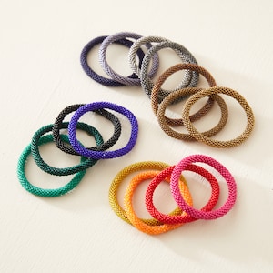 Pode incluir: Um conjunto de 14 pulseiras de contas coloridas em vários tons de azul, verde, vermelho, laranja e dourado. As pulseiras são feitas de contas pequenas e bem entrelaçadas e estão empilhadas em um padrão circular.