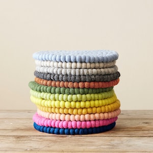 May include: A stack of circular felt ball coasters in a variety of colours including light blue, grey, charcoal, orange, green, lime, yellow, gold, peach, pink, and dark blue. Each coaster is constructed from numerous small, round felted wool balls.