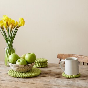 Puede incluir: Un juego de cuatro posavasos de fieltro verde con una taza de cer&aacute;mica blanca encima de uno de los posavasos. La taza est&aacute; sobre una mesa de madera con un cuenco de manzanas verdes y un jarr&oacute;n de narcisos amarillos en el fondo.