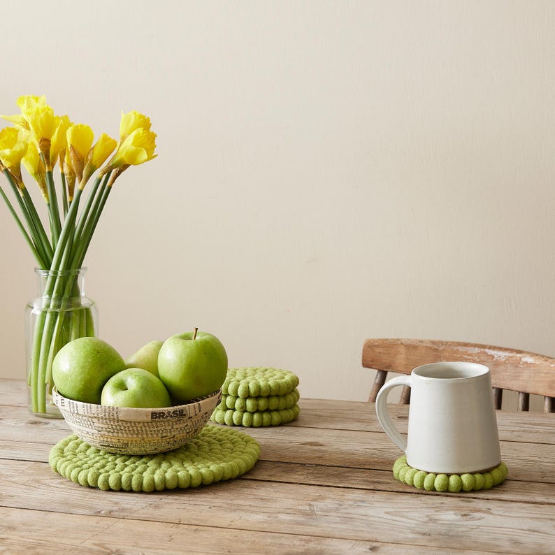 Puede incluir: Un juego de cuatro posavasos de fieltro verde con una taza de cer&aacute;mica blanca encima de uno de los posavasos. La taza est&aacute; sobre una mesa de madera con un cuenco de manzanas verdes y un jarr&oacute;n de narcisos amarillos en el fondo.