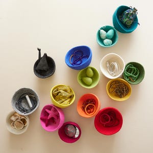 Puede incluir: Una colección de cuencos de fieltro en varios colores, cada uno lleno de diferentes artículos, como joyas, cables y objetos pequeños. Los cuencos están dispuestos sobre una superficie blanca.