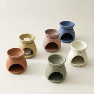Può includere: Una collezione di sei bruciatori di essenze in ceramica in vari colori opachi, tra cui terracotta, giallo senape, rosa antico, verde salvia, crema e blu ardesia. Ogni bruciatore presenta una base arrotondata con un'ampia apertura e un piattino.