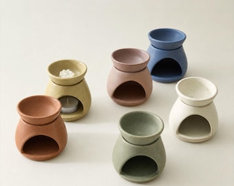May include: A collection of six ceramic oil burners in various matte colors including terracotta, mustard yellow, dusty rose, sage green, cream, and slate blue. Each burner features a rounded base with a wide opening and a removable top dish.