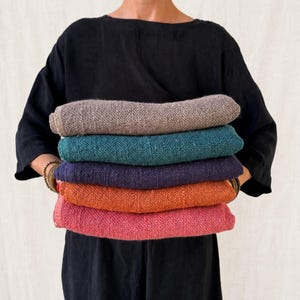 May include: A stack of five folded rectangular textiles with a visible coarse weave. The colours from top to bottom are light brown, teal, dark purple, burnt orange, and bright pink. Each piece of fabric has a consistent, textured appearance.