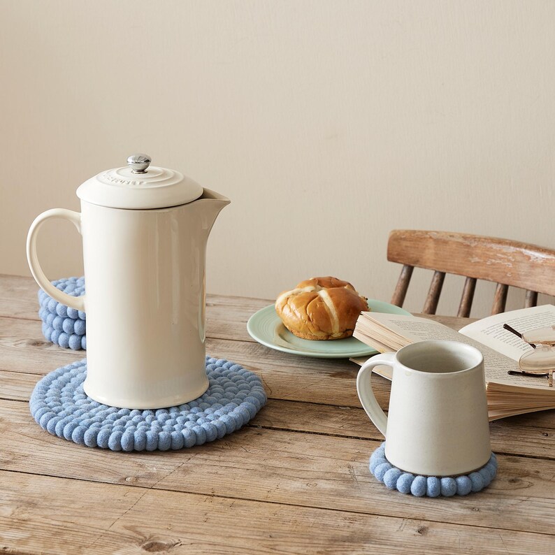 Puede incluir: Una cafetera de prensa francesa de cer&aacute;mica blanca sobre un posavasos de punto azul. Una taza de cer&aacute;mica blanca sobre un posavasos de punto azul. Un libro y un plato con un pastel sobre una mesa de madera.