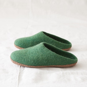 May include: A pair of forest green felted wool slippers with a rounded toe and an open back design. The slippers feature a textured, fuzzy surface and are constructed with a thin, flat brown sole that provides a base for the soft upper material.
