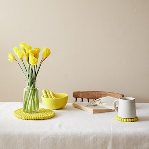 Puede incluir: Una taza de cer&aacute;mica blanca con un posavasos de fieltro amarillo se encuentra sobre un mantel blanco junto a un libro y un cuenco de fieltro amarillo. Un jarr&oacute;n con narcisos amarillos se encuentra sobre la mesa.