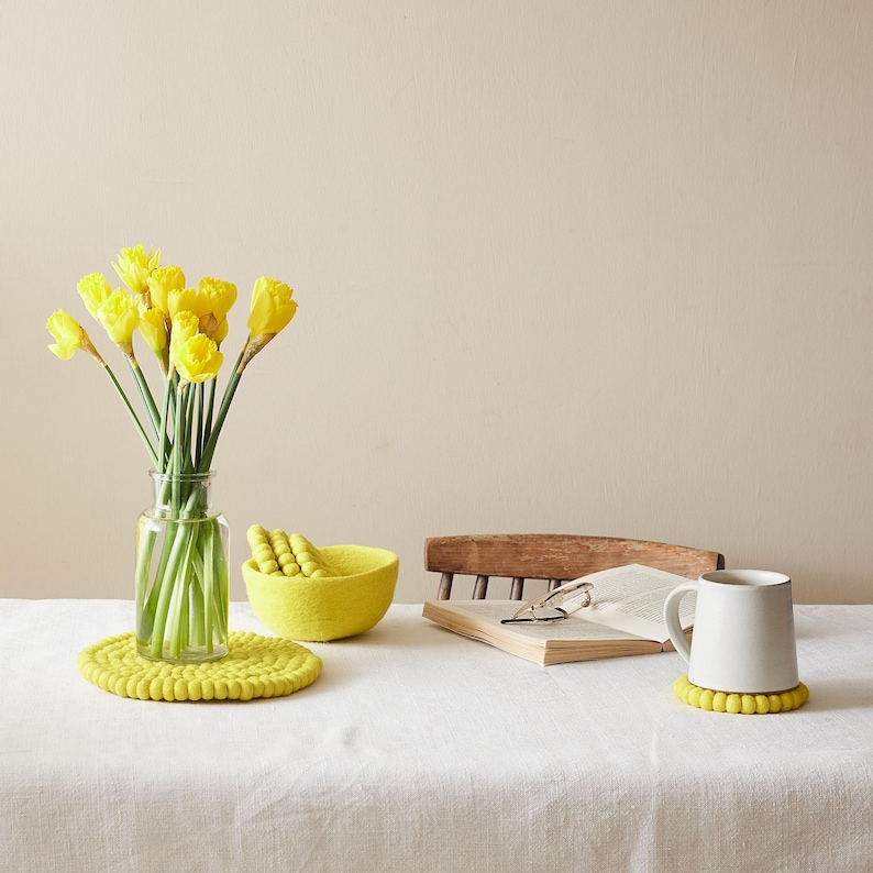 Puede incluir: Una taza de cer&aacute;mica blanca con un posavasos de fieltro amarillo se encuentra sobre un mantel blanco junto a un libro y un cuenco de fieltro amarillo. Un jarr&oacute;n con narcisos amarillos se encuentra sobre la mesa.
