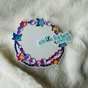 Peut inclure: Un autocollant blanc avec un motif circulaire représentant des formes de renard et d'étoile bleues, des perles violettes et le mot "BLUE". L'autocollant comprend également le texte "stella forbes energy".