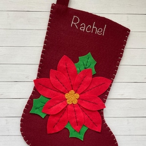 May include: A burgundy felt Christmas stocking featuring a red poinsettia flower and green leaves. The name "Rachel" is embroidered in white thread. White stitching outlines the stocking, a festive holiday decoration.