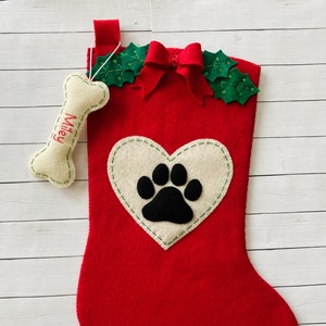 May include: Red felt Christmas stocking with a white heart and black paw print design. The stocking has a green holly and red bow at the top and a white bone ornament with the name "Miley" embroidered on it.