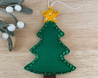 May include: A green felt Christmas tree ornament with a yellow star on top, embellished with sequins. The tree has a brown trunk and a white string for hanging. A festive holiday decoration.
