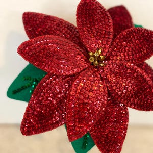 Könnte beinhalten: Eine rote Weihnachtsstern-Blume aus Pailletten mit grünen Blättern. Die Blume ist eine Weihnachtsdekoration.