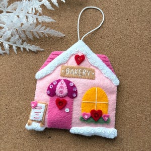 Puede incluir: Un adorno de panadería de fieltro rosa con ribete blanco y una anilla para colgar. El adorno presenta una puerta, una ventana y un letrero que dice "BAKERY". Corazones y botones decorativos añaden detalles.