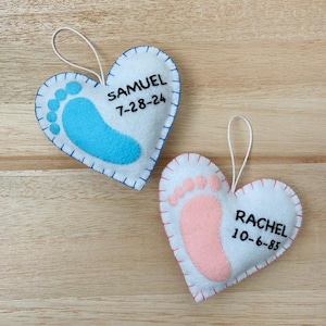 May include: Two white felt heart ornaments with blue and pink footprints and embroidered names and dates. The blue ornament says "Samuel 7-28-24" and the pink ornament says "Rachel 10-6-85".