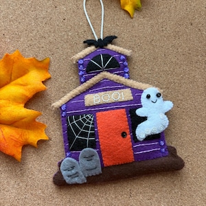 May include: A purple felt house ornament with an orange door, a white ghost, and two gray tombstones. The house features a black spiderweb window, a black bat on the roof, and the word BOO! stitched in white thread. It includes a white hanging loop.