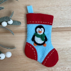 May include: A small blue felt stocking with a red cuff and a penguin applique. The penguin is wearing a green scarf and has a white belly and orange feet.