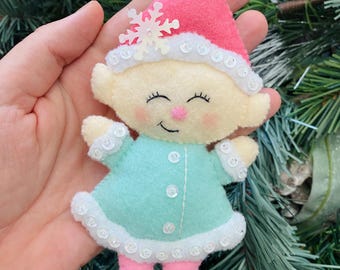 Könnte beinhalten: Handgefertigter Weihnachtsbaumschmuck aus Filz in Form eines Elfen. Der Elf hat einen rosa Hut, einen hellblauen Mantel, rosa Leggings und blaue Stiefel. Der Schmuck ist mit weißen Pailletten und einem Schneeflocken verziert.