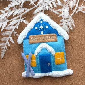 May include: A blue felt ski lodge ornament with white trim and a snowflake design. The ornament has a door, two yellow windows, and the words "SKI LODGE" on a brown banner. A pair of blue skis is attached to the side.