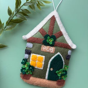 May include: A felt ornament shaped like a pub, featuring a green roof with brown accents and a white trim. The facade is olive green with a door, window, and the word "PUB". Decorative shamrocks and flower pots add detail. A white string is attached for hanging.