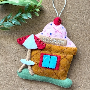Puede incluir: Un adorno de fieltro con forma de heladería con base de cono de barquillo, techo de helado rosa y sombrilla roja y blanca. El letrero dice ICE CREAM en hilo blanco. Cuenta con una ventana azul, una cereza roja y un lazo blanco para colgar.