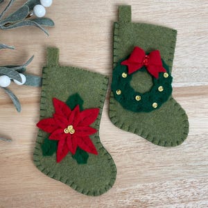 Può includere: Due calze di Natale in feltro verde con decorazioni festive. Una calza presenta una stella di Natale rossa con accenti di glitter dorati, mentre l'altra ha una ghirlanda verde con un fiocco rosso e accenti di glitter dorati.