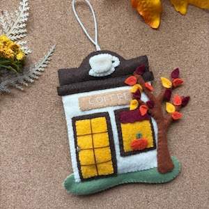 May include: A felt hanging ornament shaped like a coffee shop with a white wall, brown roof, and yellow door. It features a white coffee cup icon, the word COFFEE embroidered in white, and a brown tree with red and yellow leaves and a small orange pumpkin.