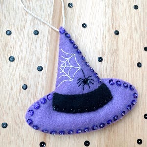 May include: Purple felt witch hat ornament with a black felt brim and white embroidered spider web and spider. The hat is decorated with purple sequins.