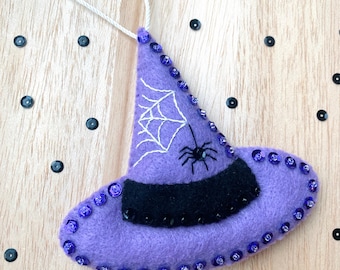 May include: Purple felt witch hat ornament with a black felt brim and white embroidered spider web and spider. The hat is decorated with purple sequins.