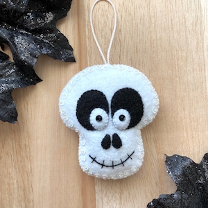 May include: White felt skull ornament with black eyes and a stitched smile. The ornament has a white string loop for hanging.