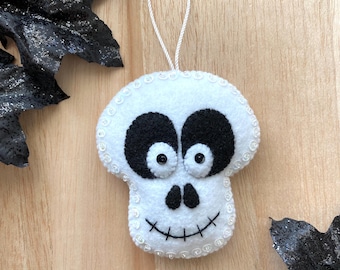 May include: White felt skull ornament with black eyes and a stitched smile. The ornament has a white string loop for hanging.